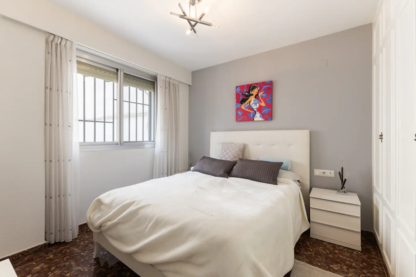 Bedroom with white bed, gray pillows, and a red painting on the gray wall. A white nightstand is next to the bed. Window with white curtains.