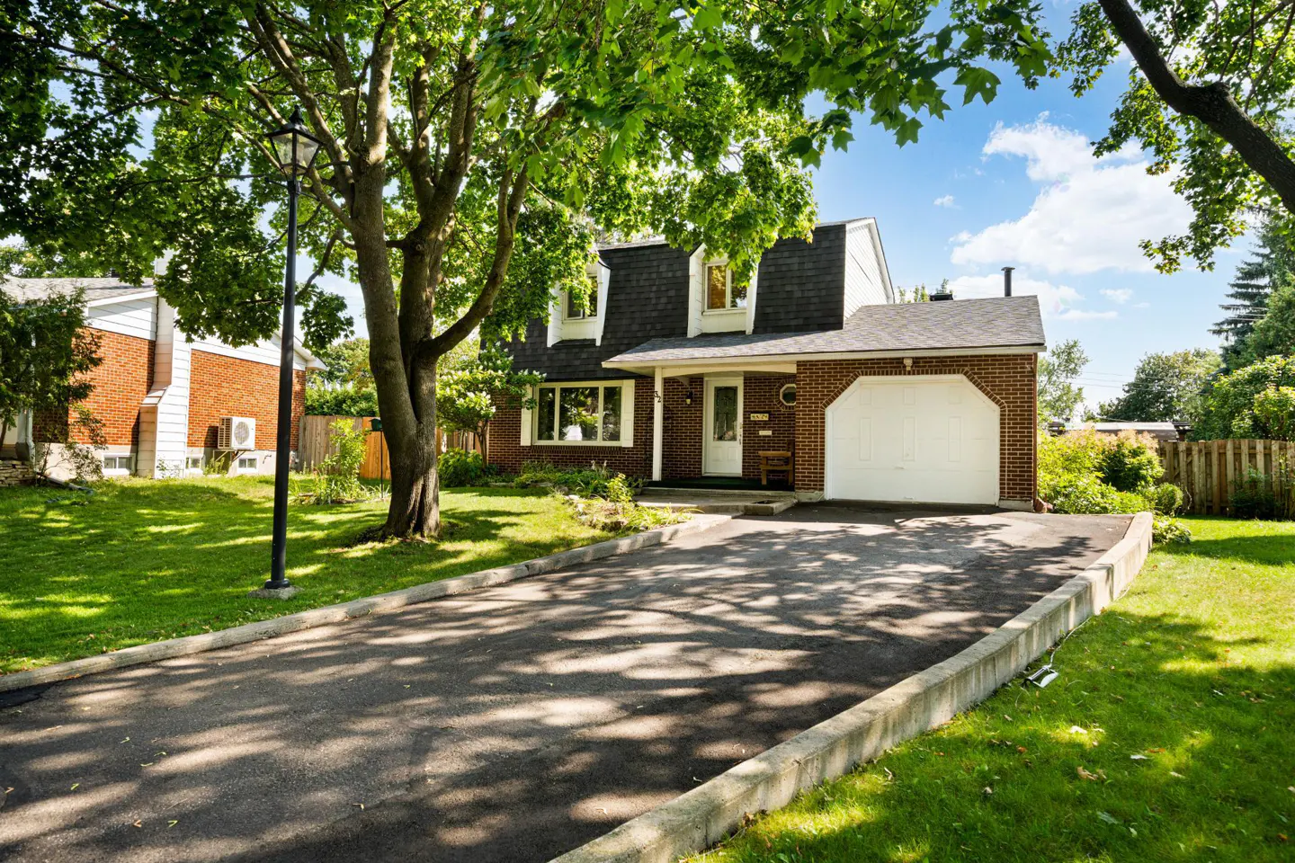 A two-story brick house with a black roof and white garage door on a sunny day. A long driveway leads to the house.