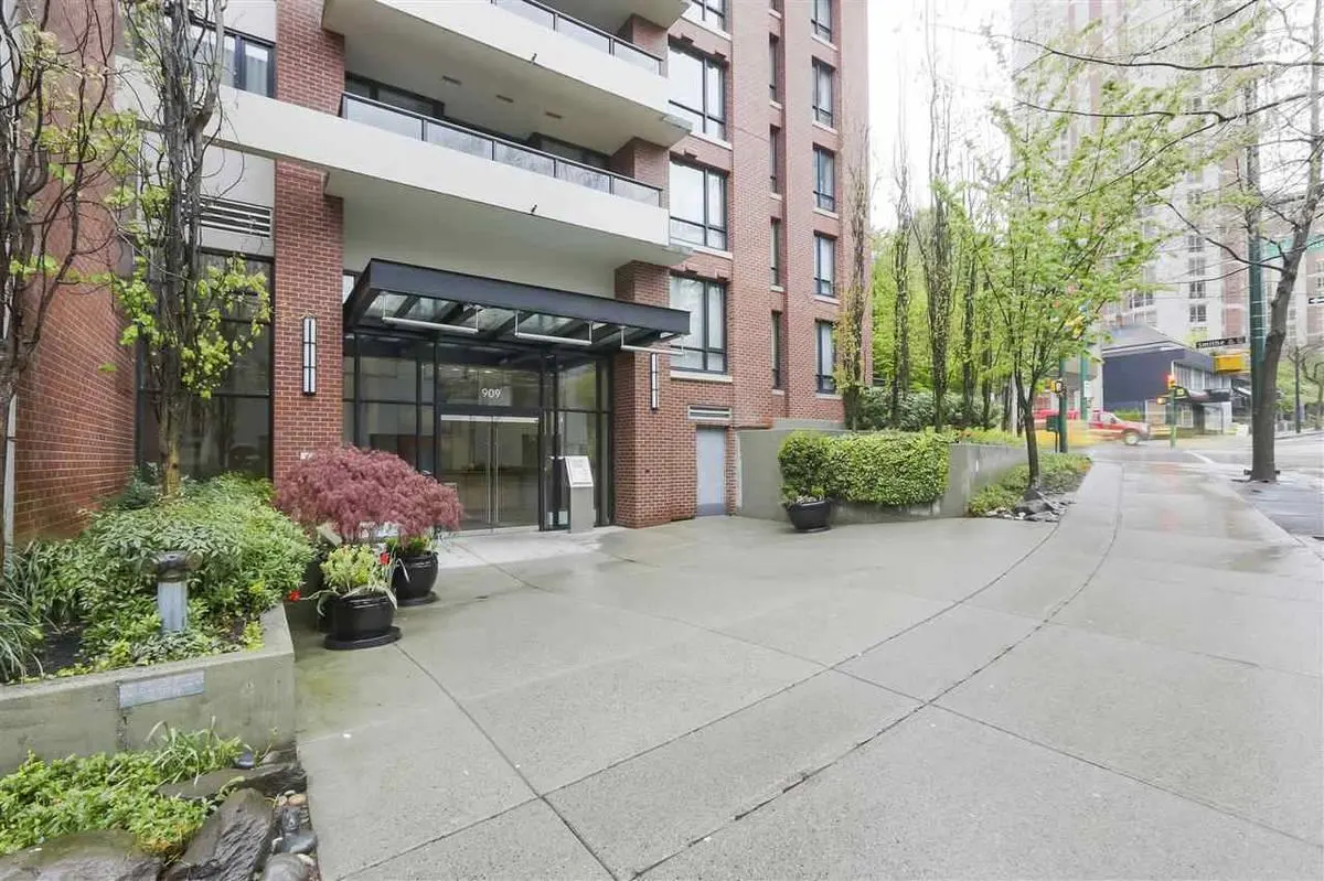 Apartment building entrance with glass doors, black awning, and brick facade. Potted plants and greenery surround the entrance.