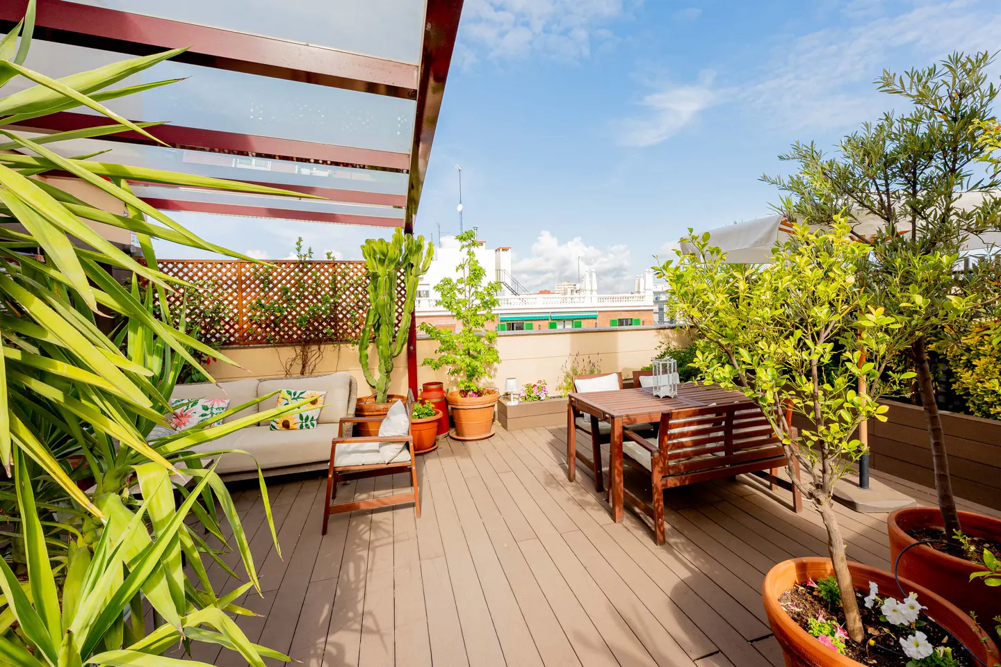 A rooftop patio with a wooden deck, a dining table, a sofa, and potted plants under a clear blue sky.