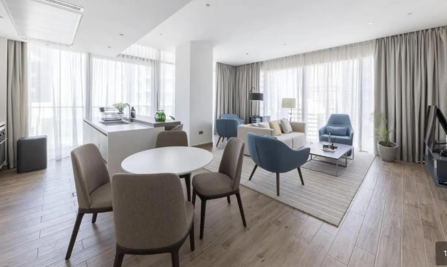 Bright, open-concept apartment with a white kitchen island, round dining table with four chairs, and a living room with blue armchairs and a beige sofa. Large windows with sheer curtains.