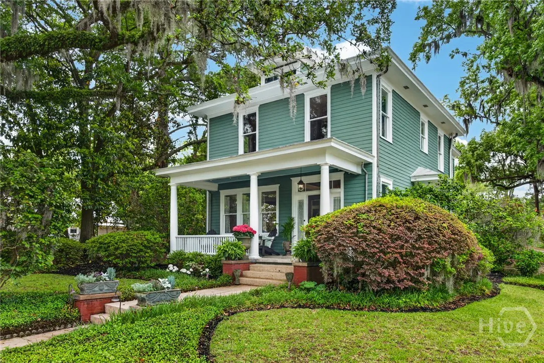 Two-story teal house with white trim and a porch with white columns. Lush green lawn and landscaping surround the home.