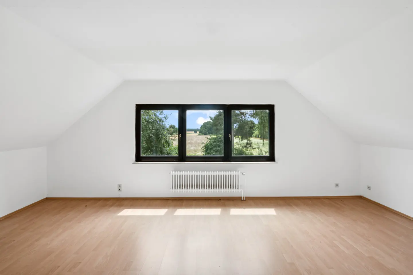 Attic room with white walls, wood floor, and black-framed window showing a field and trees. A white radiator sits below the window.