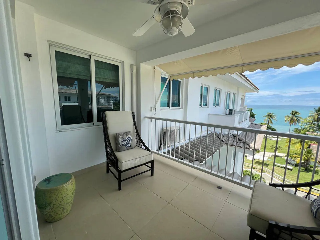 Balcony with chairs overlooking the ocean. White building with awning, ceiling fan, and green side table.