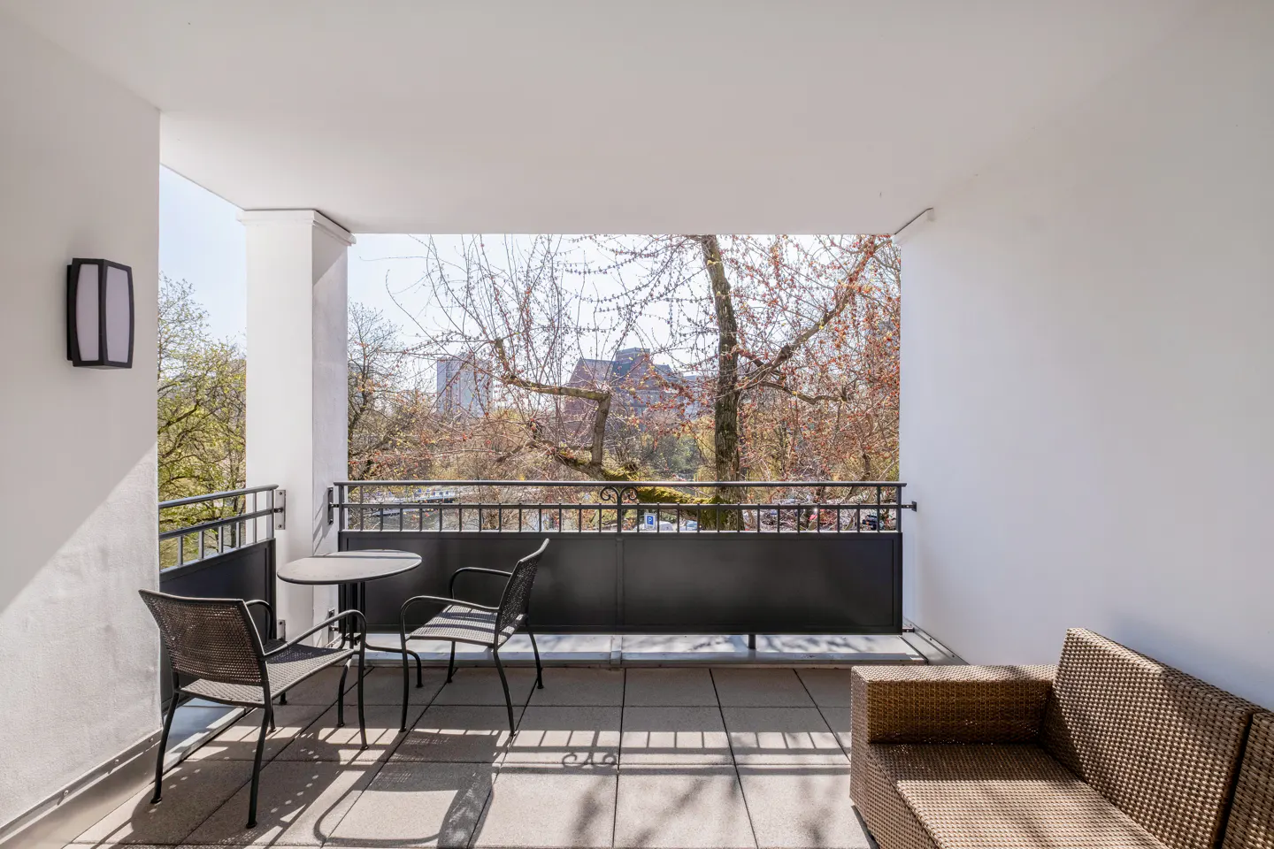 A bright balcony with a table, chairs, and a wicker couch overlooks trees and a city.