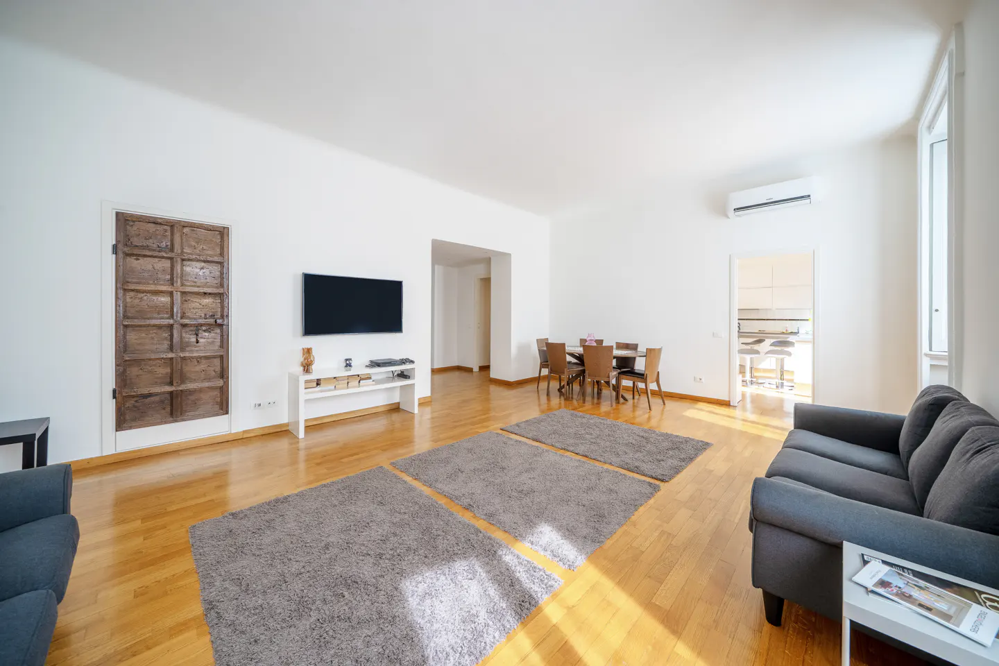 Bright living room with hardwood floors, grey sofa, three rugs, wall-mounted TV, and a dining area in the background.