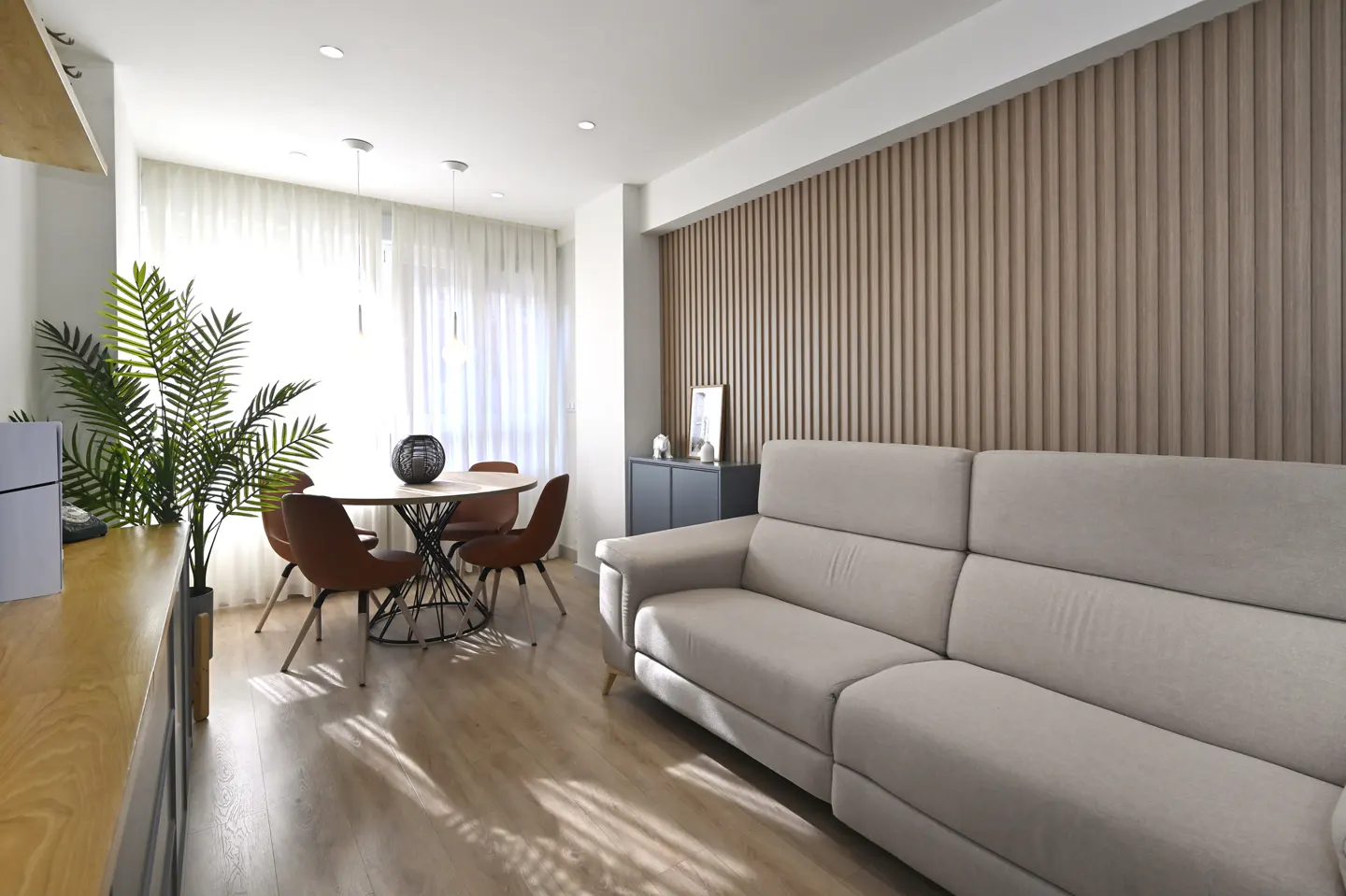 Bright living room with a beige sofa, round table with four chairs, and a wood-paneled wall. A plant and cabinet are also visible.