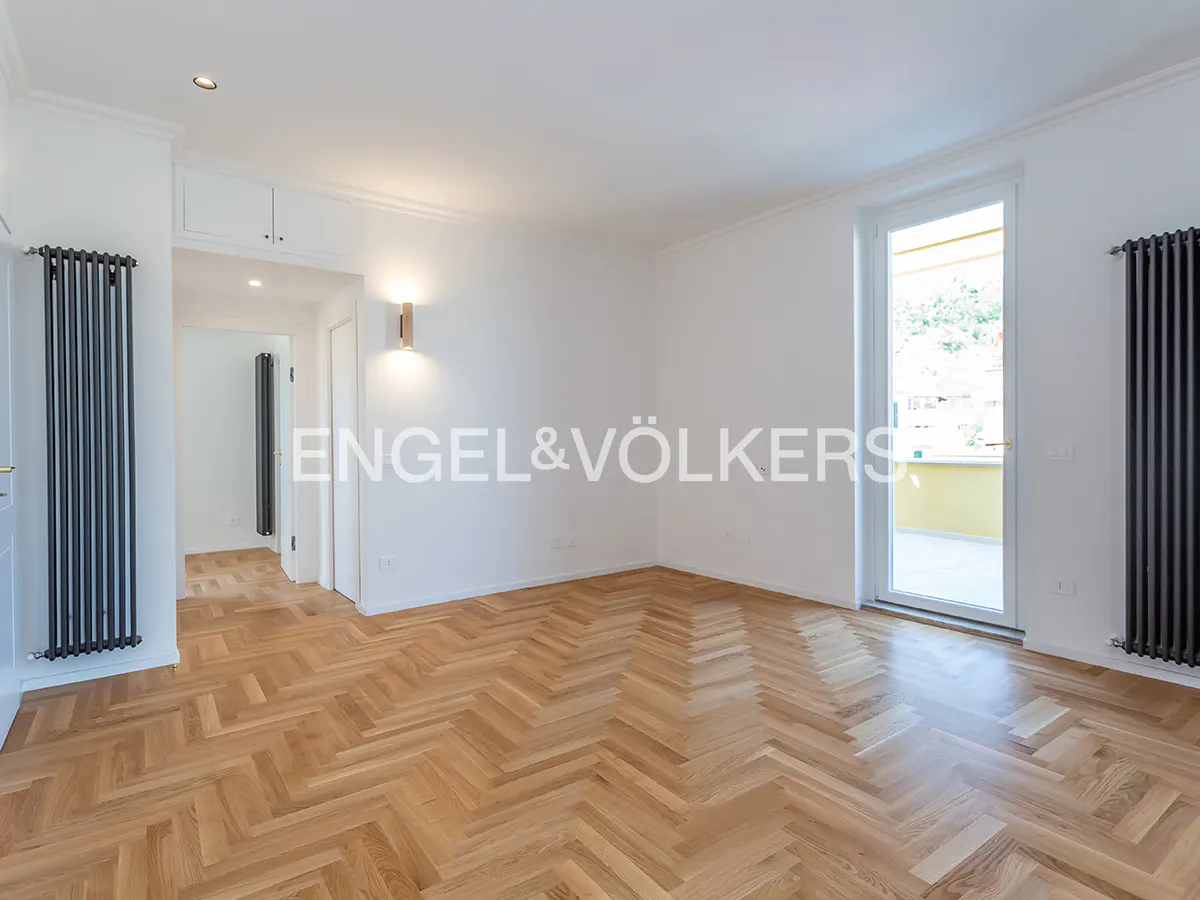Bright, empty room with herringbone wood floors, white walls, and a balcony view. Engel & Völkers logo is visible.