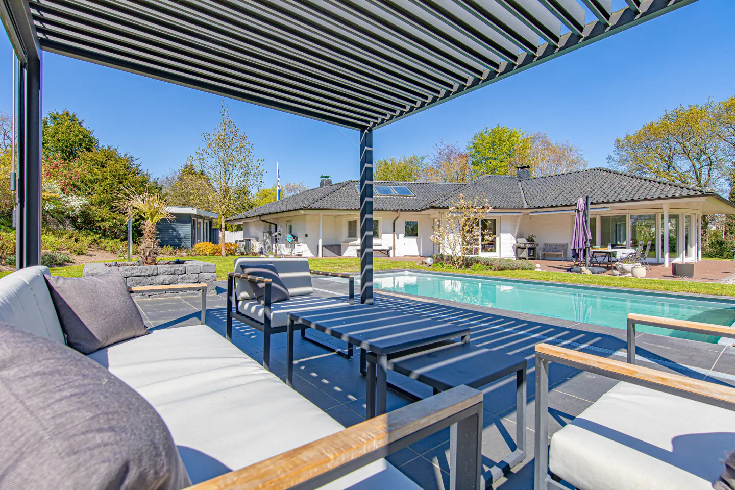 Outdoor patio with white cushioned seating under a pergola, overlooking a pool and a white house with a black roof.