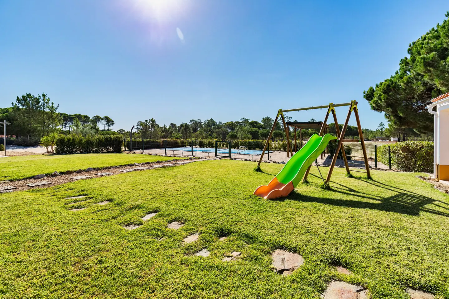 A sunny backyard with a green and orange slide, swing set, pool, and stone path on a lush lawn.