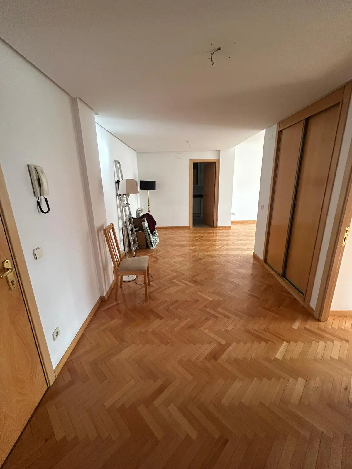 Bright, empty room with herringbone wood floors, white walls, and a wooden chair. A ladder and furniture are in the background.