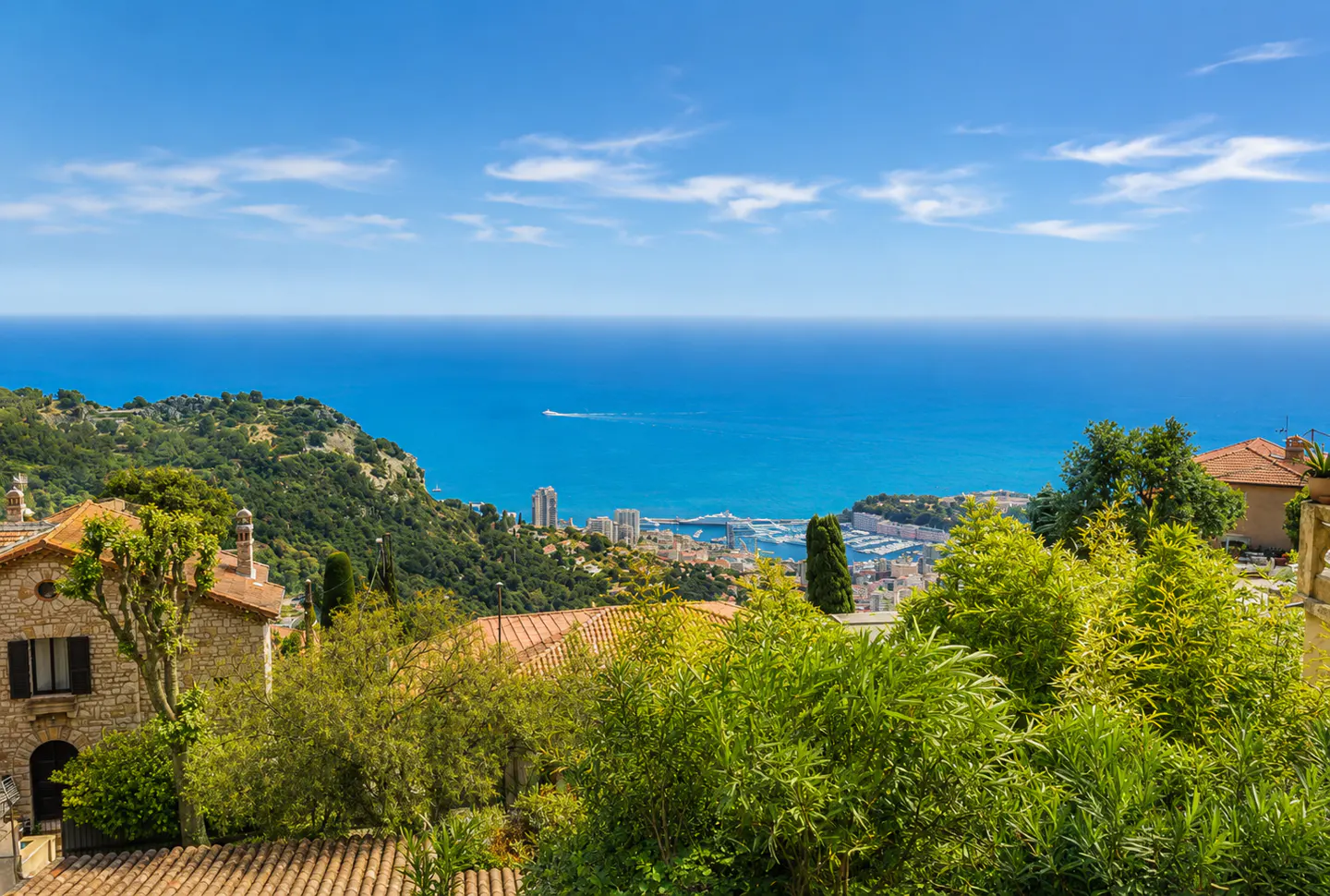 Scenic view of Monaco with stone houses, green trees, and the blue Mediterranean Sea under a clear sky.