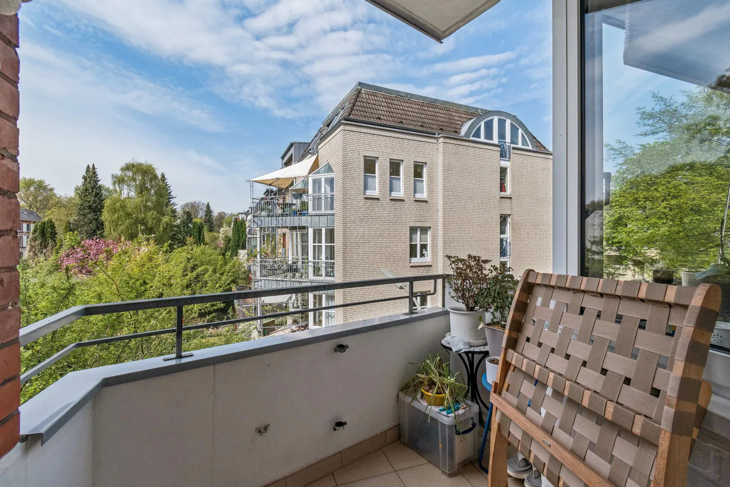 Balcony view with a woven chair, plants, and a view of a brick building and green trees under a blue sky.