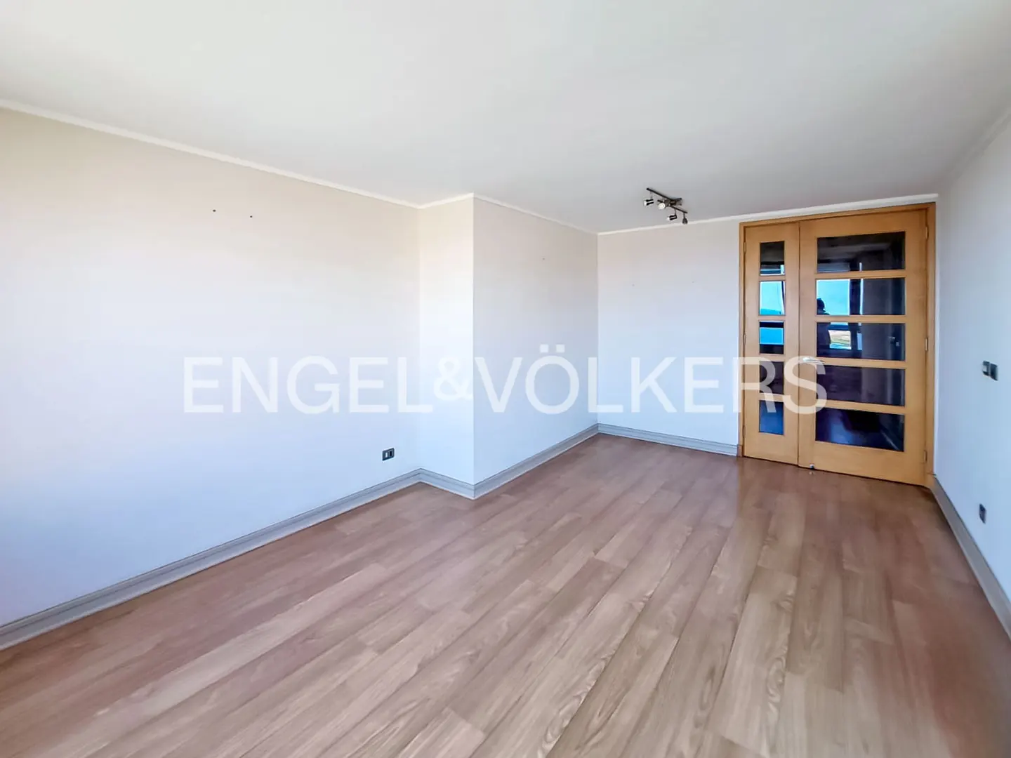 Empty room with light wood floors, white walls, and a sliding glass door to a balcony with an ocean view. Engel & Völkers logo on the wall.