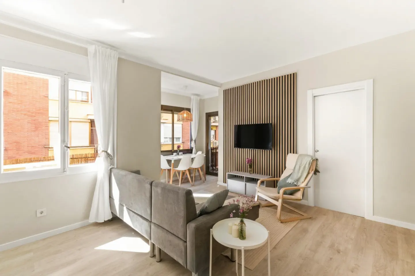 Bright living room with a gray sofa, wood accent wall with a TV, and a dining area visible in the background.