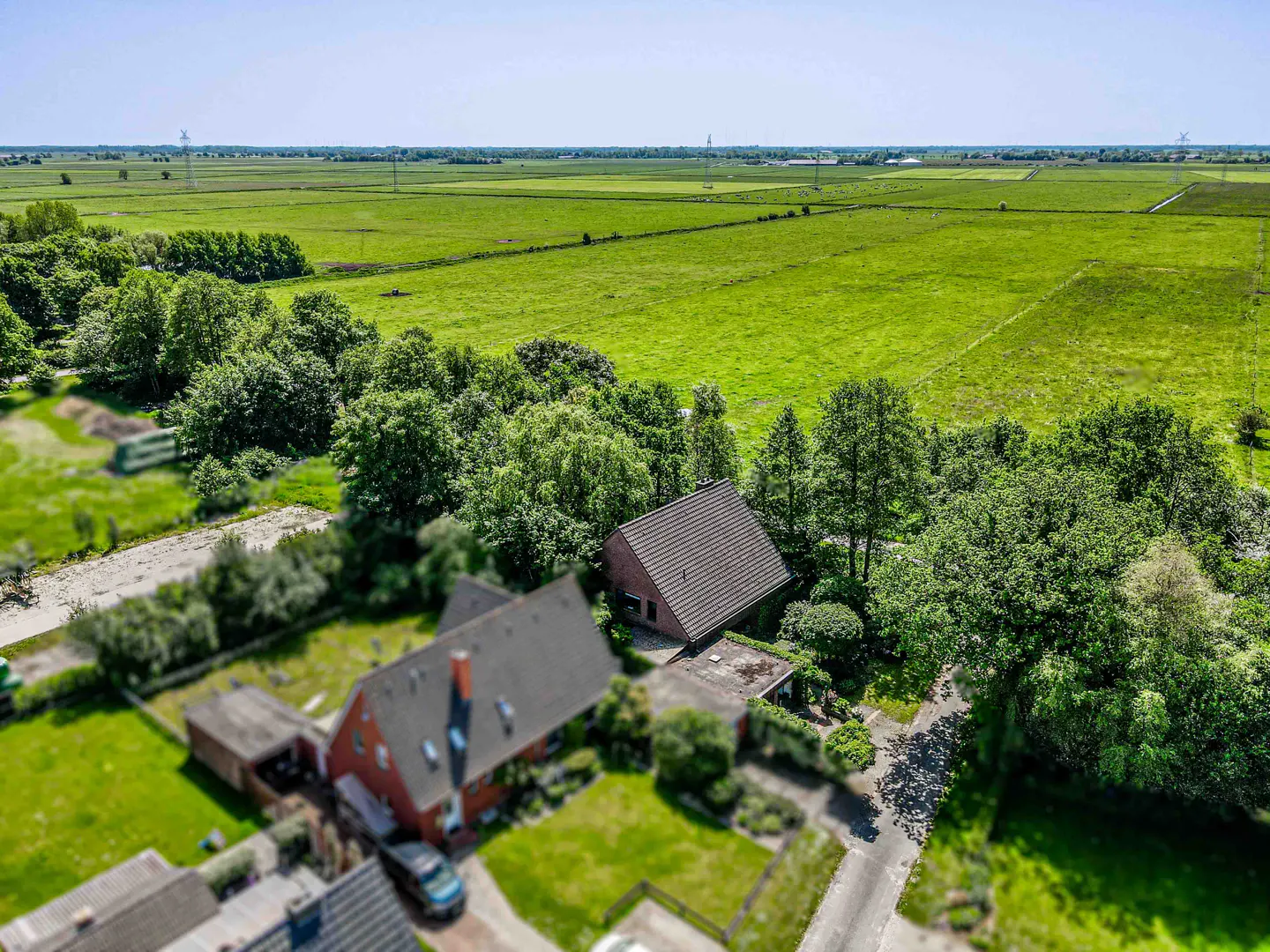 Aerial view of a red brick house surrounded by lush green trees and expansive flat farmland under a clear blue sky.