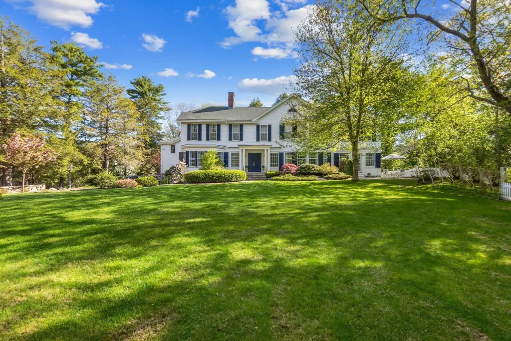 A white colonial-style house with blue shutters sits on a large green lawn under a blue sky with scattered clouds.
