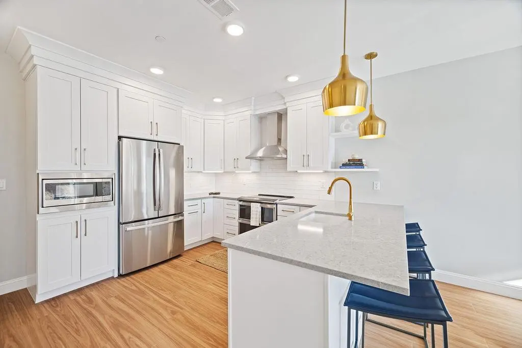 Bright kitchen with white cabinets, stainless steel appliances, and wood floors. Gold pendant lights hang over a white countertop island with blue stools.
