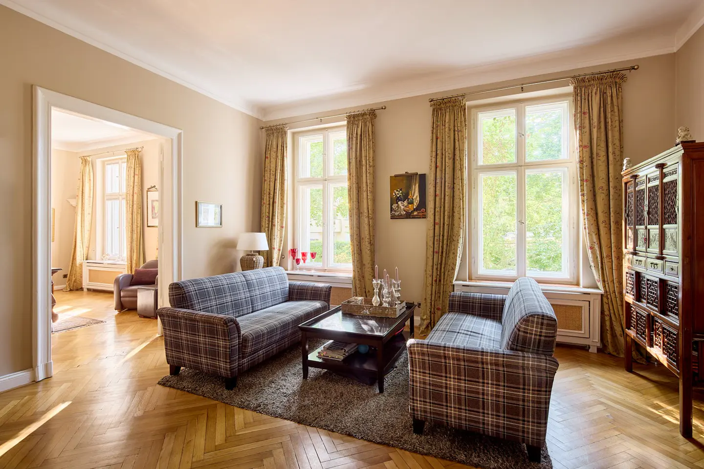 Living room with two plaid sofas, a dark wood coffee table, and a dark wood cabinet. Two windows with floral curtains let in natural light.