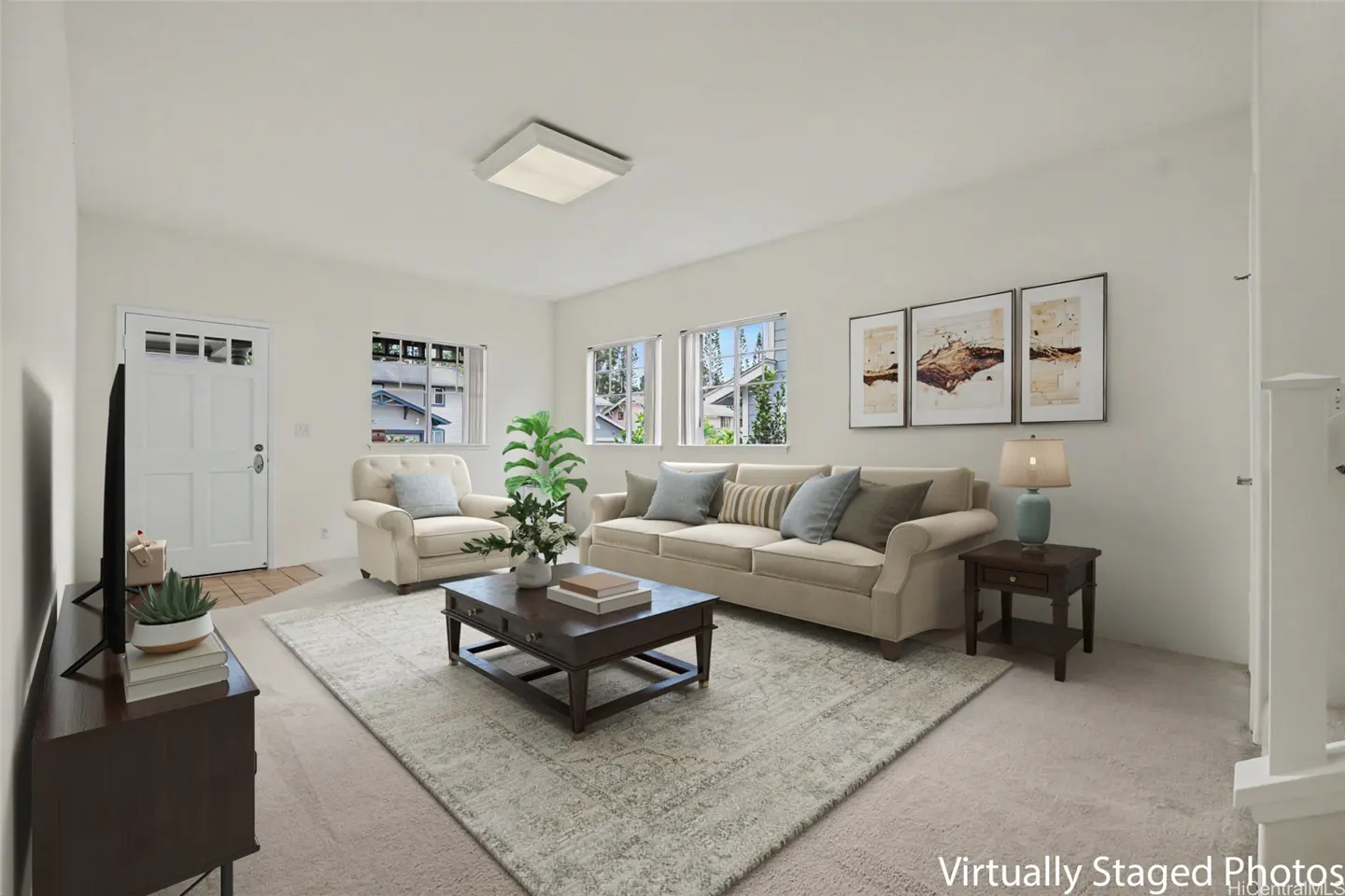 Bright living room with beige sofa, chair, and patterned rug. Dark wood coffee table and side table with lamp. White walls and door.