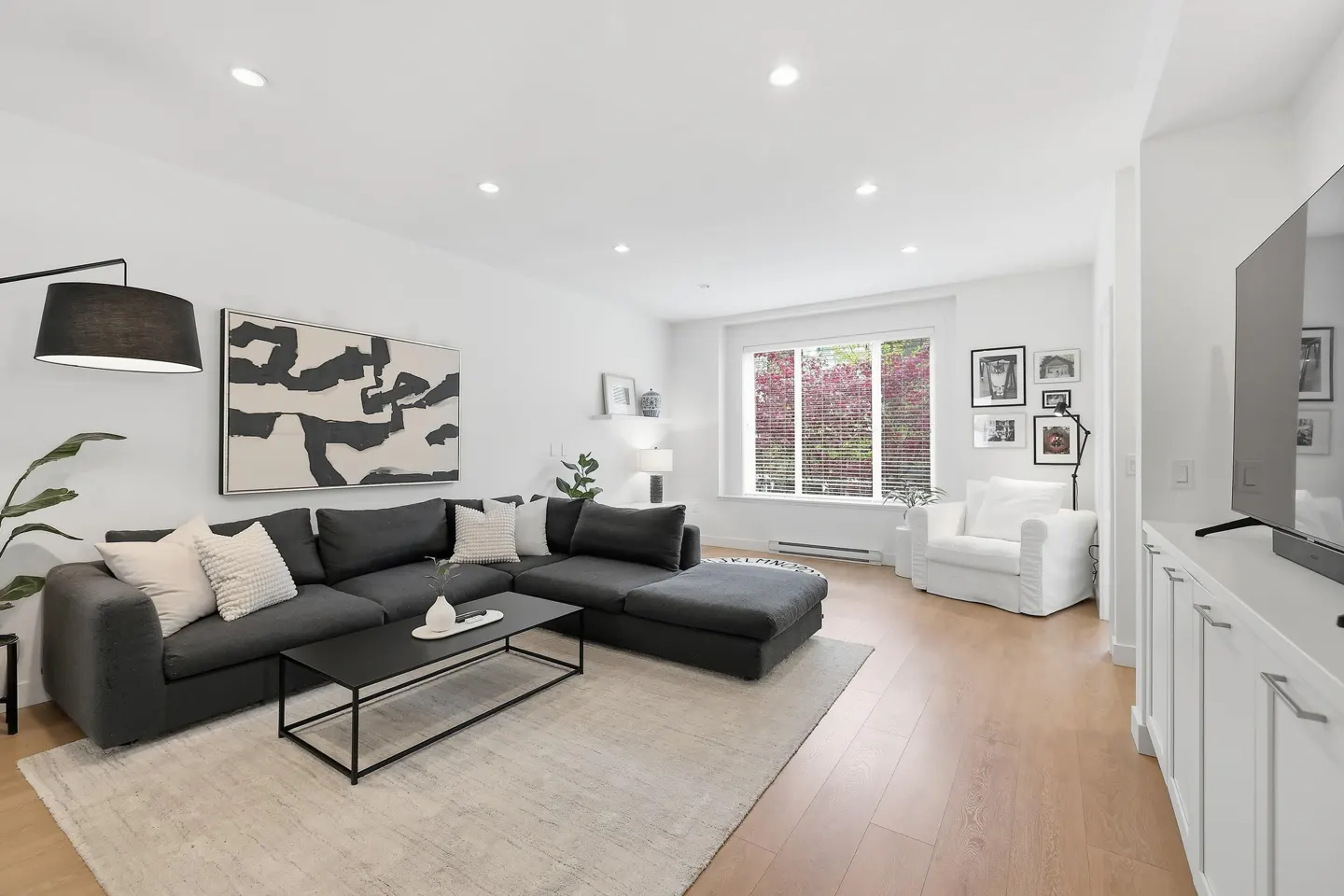 Bright living room with hardwood floors, a gray sectional sofa, a black coffee table, and a large abstract painting on the white wall.