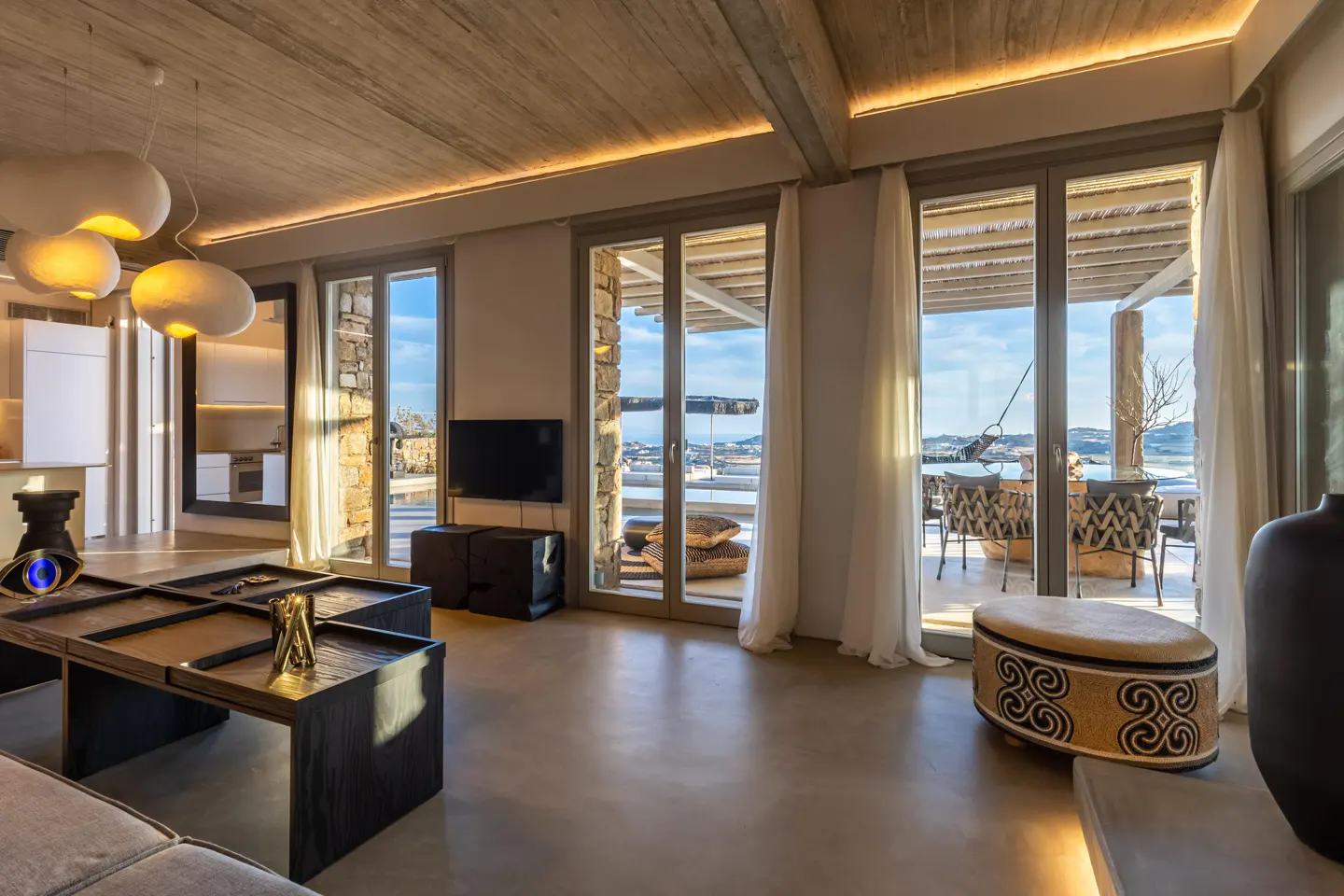 A modern living room with a wooden ceiling, pendant lights, and a view of the ocean through large glass doors.