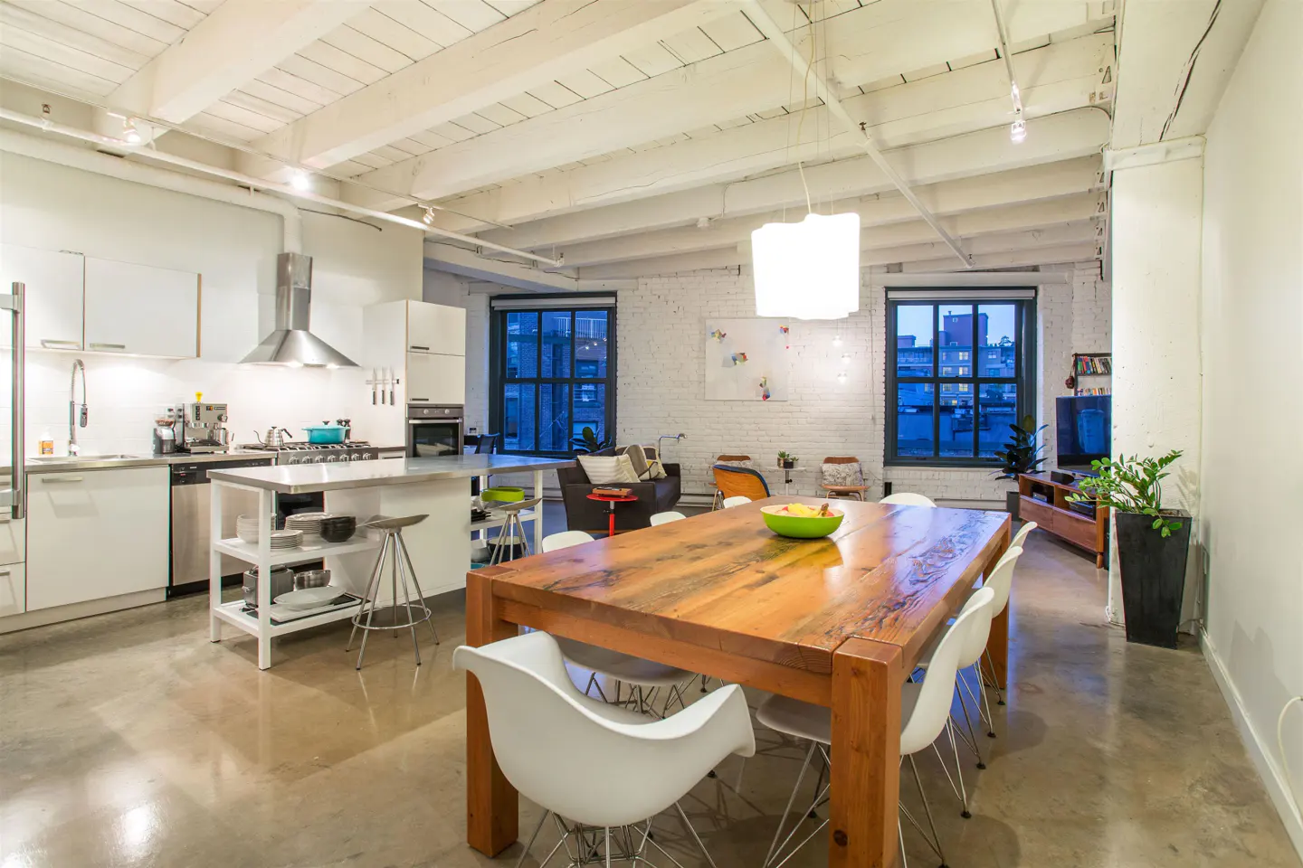 Bright, open loft with a white kitchen, stainless steel appliances, and a large wooden dining table with white chairs. Windows overlook a city view.
