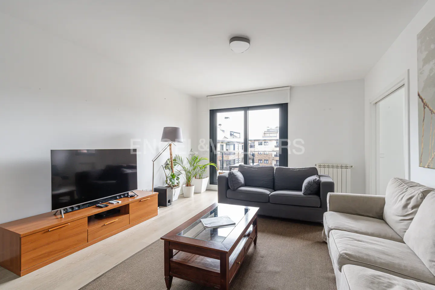 Bright living room with a gray sofa, wooden TV stand, and a glass-topped coffee table on a brown rug. A large window offers a city view.