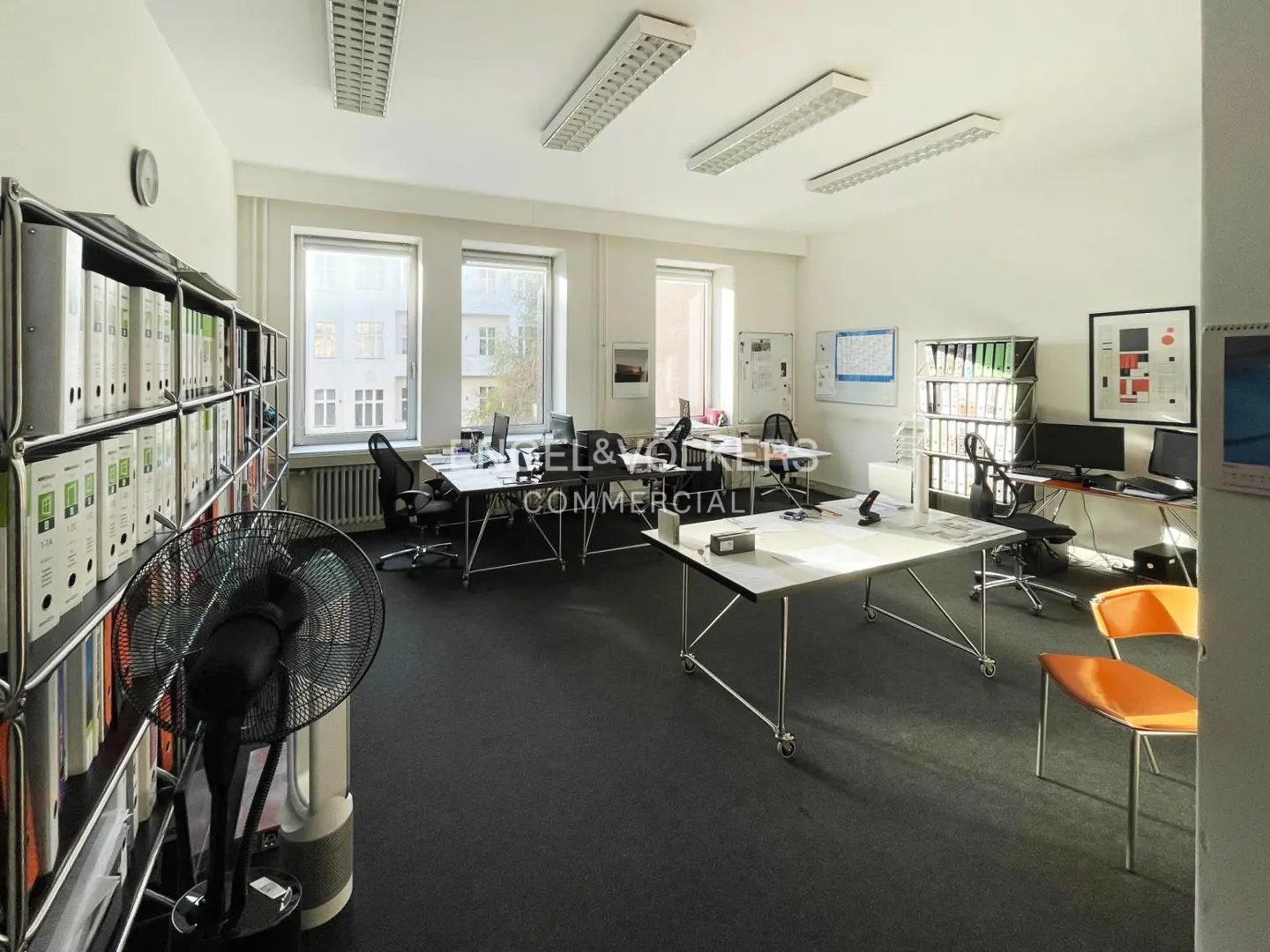 Bright office space with white walls, black carpet, and white desks. Shelves filled with binders line one wall, and windows offer natural light.