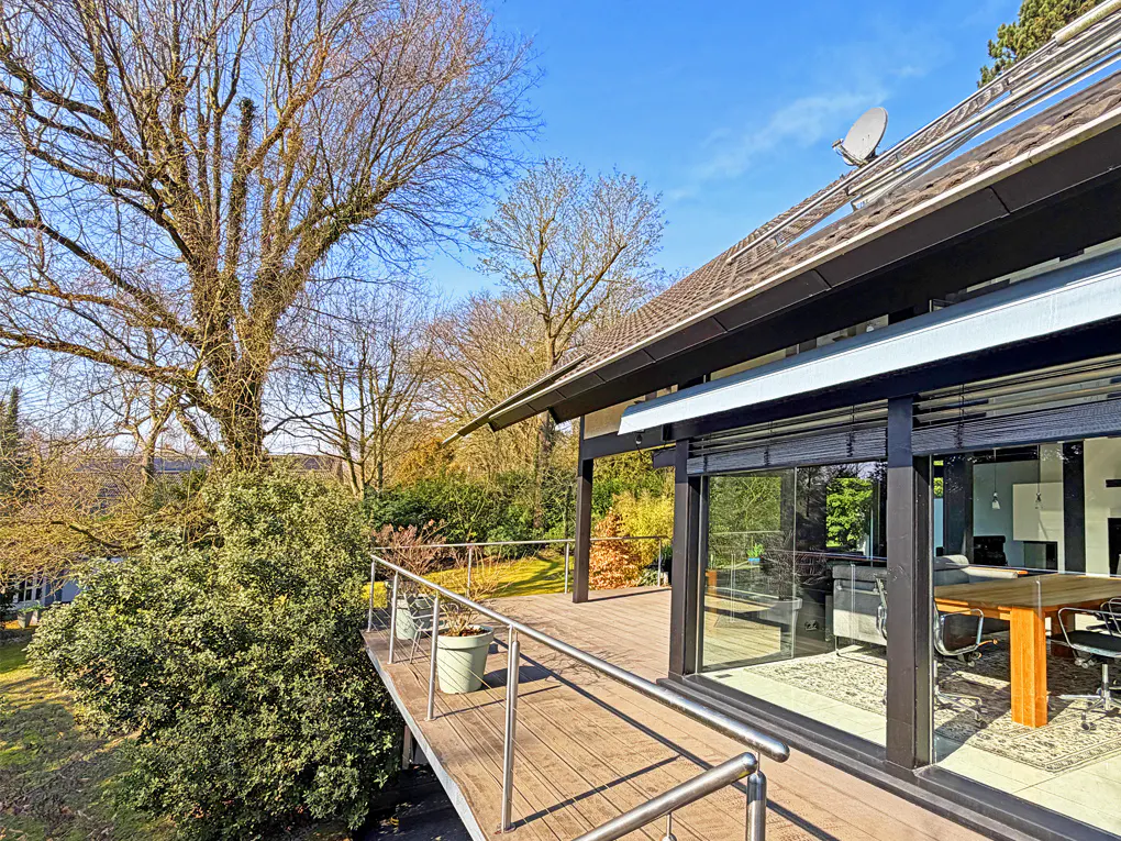 Exterior view of a modern home with a wooden deck, metal railing, and sliding glass doors. Trees and blue sky in the background.