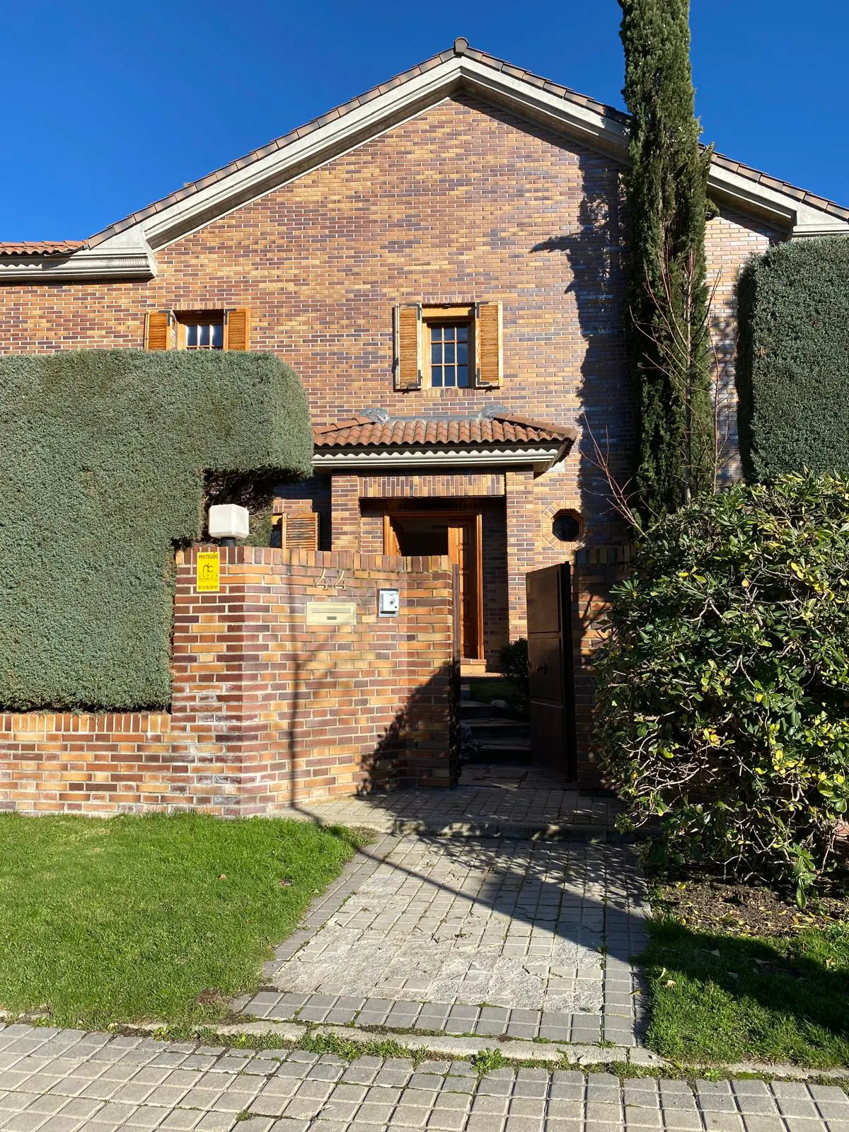 Brick house with brown shutters and a red tile roof. A green hedge and tree flank the entrance. A stone walkway leads to the front door.