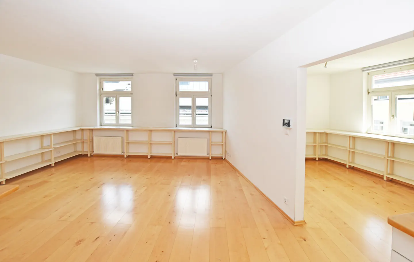 Bright, empty room with light wood floors, white walls, and built-in shelving under two windows. Another room is visible through an opening.