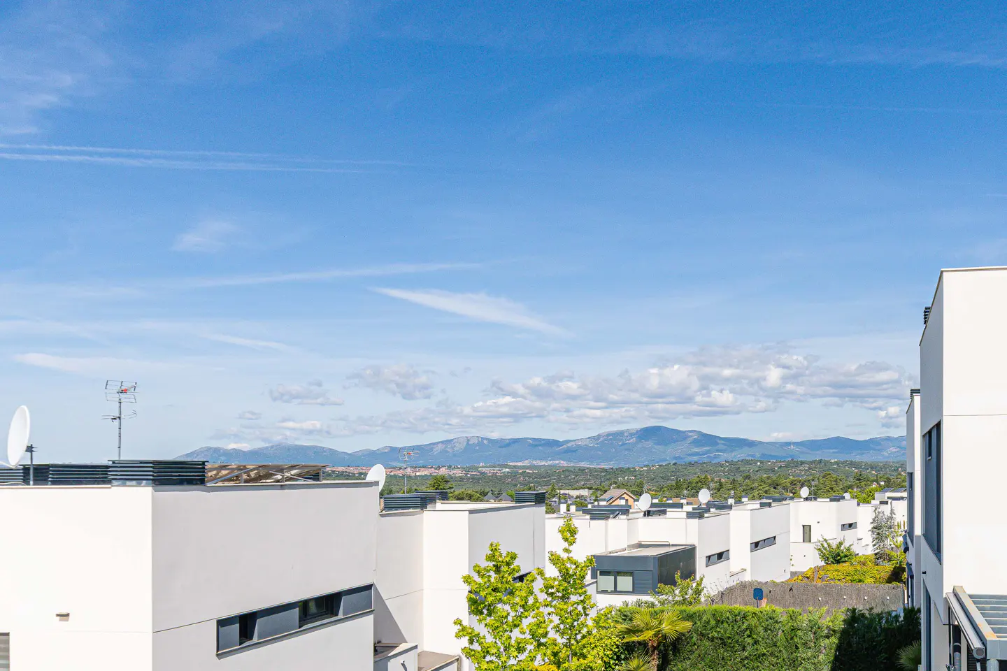 View of modern white houses under a blue sky with mountains in the distance. Trees and green hedges are visible.