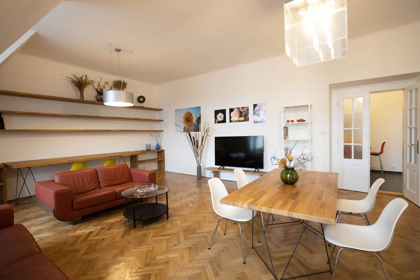 Bright living room with wood floors, red leather sofa, and a wood table with white chairs. Shelves line the wall above the sofa.