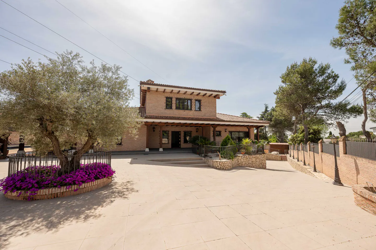 Two-story brick house with a red tile roof and a large paved front yard. A tree with purple flowers sits in front.