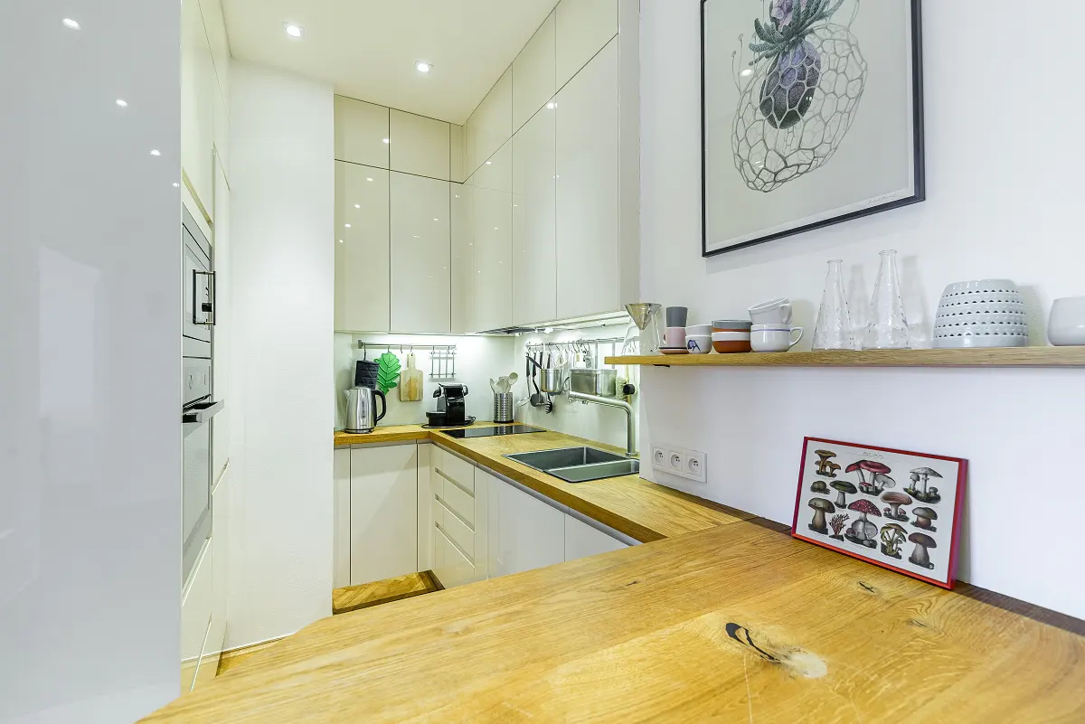 Bright kitchen with white cabinets, wood countertops, and stainless steel appliances. A shelf displays glassware and art hangs on the white walls.