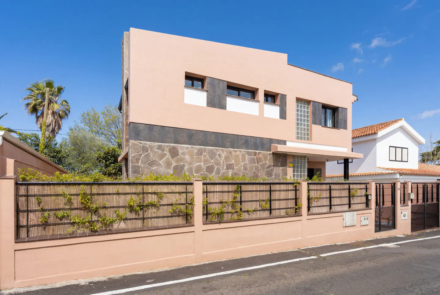 Two-story peach house with stone and dark gray accents, a bamboo fence with vines, and a blue sky.