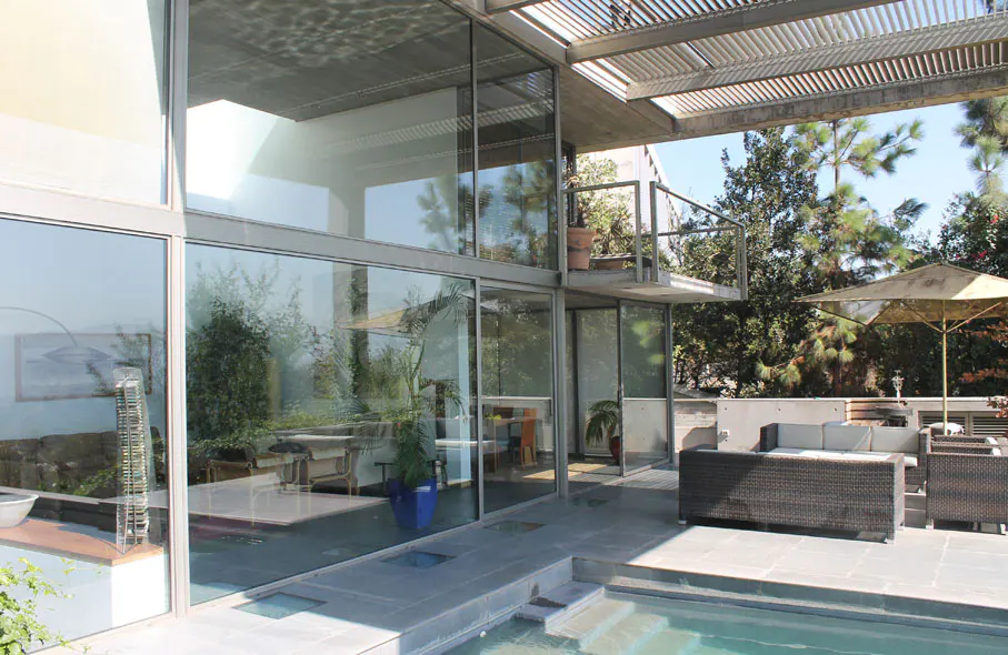 Modern home exterior with floor-to-ceiling windows, a patio with wicker furniture, and a pool. Trees are visible in the background.
