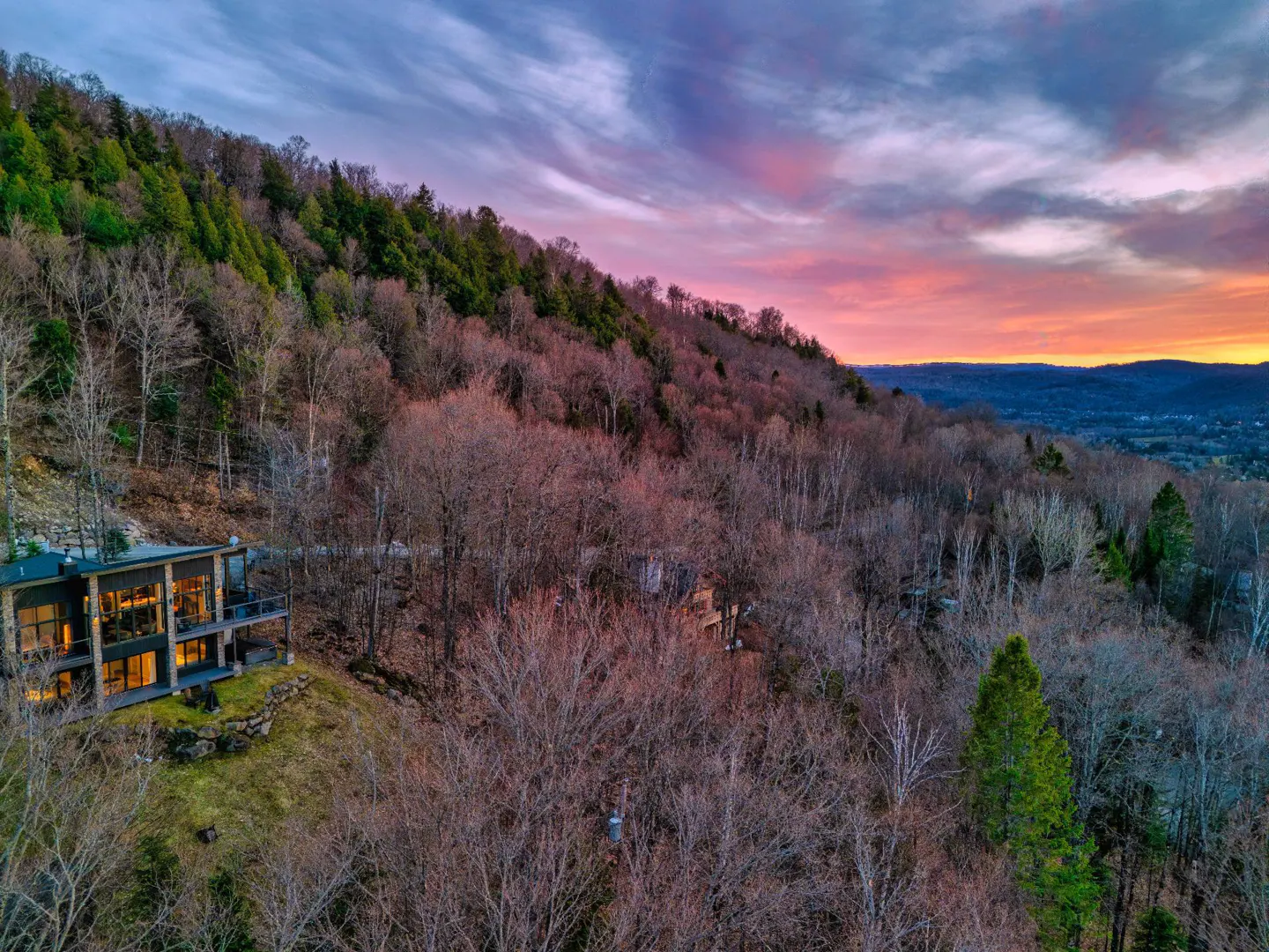Modern home on a wooded hillside at sunset. The sky is pink and orange. The house has large windows and a balcony.