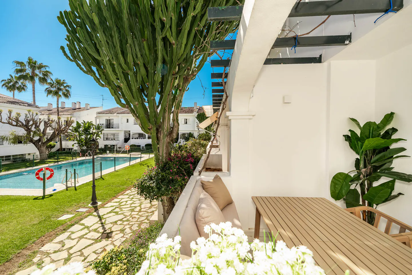 Balcony view of a pool and white buildings. A large cactus and flowers are visible. A wooden table and cushioned seating are on the balcony.