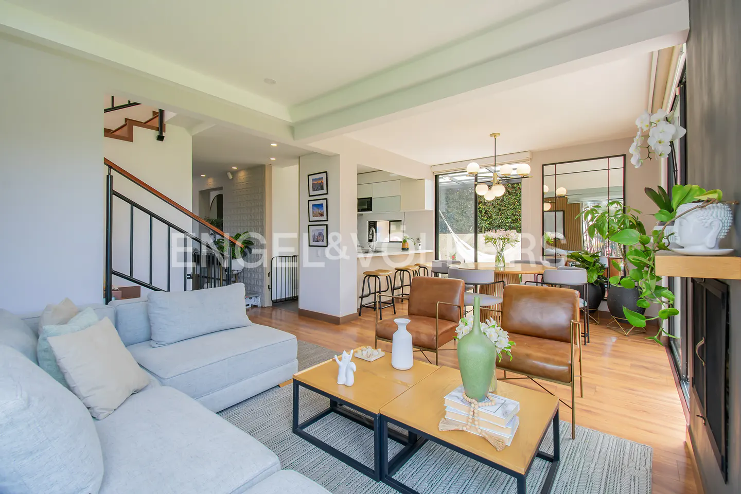 Bright, open-concept living space with hardwood floors, a gray sectional sofa, and leather chairs. A modern kitchen and dining area are visible in the background.