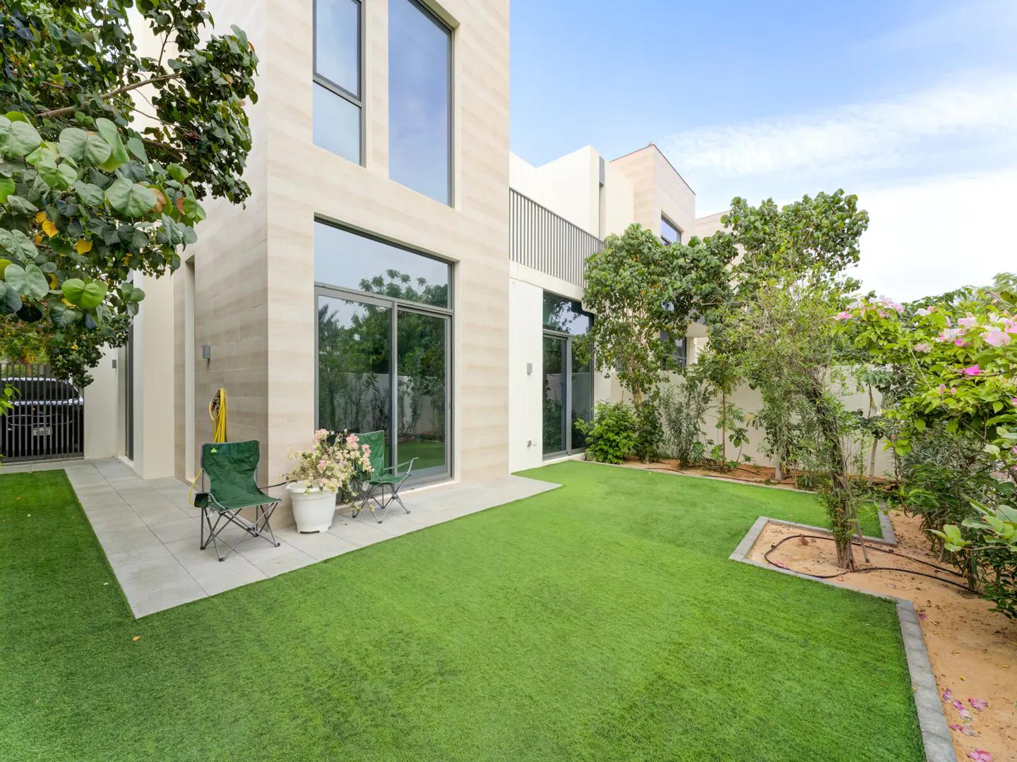 Modern home exterior with green lawn, patio, and chairs. Large windows reflect trees. Flowers in a pot sit between the chairs.