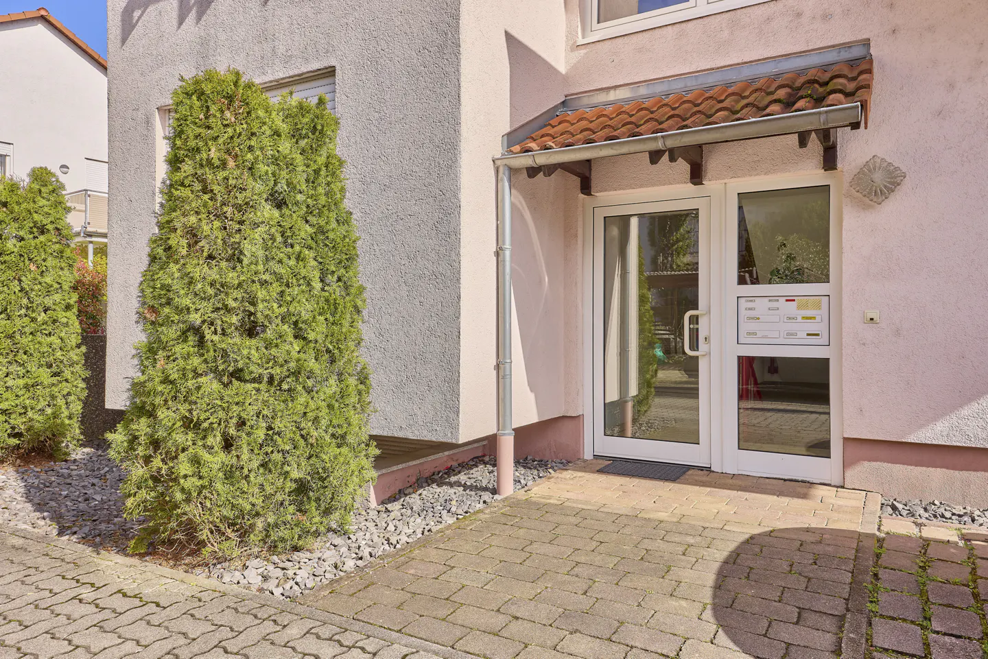 Exterior view of a pink building with a white door and red tiled awning. Green shrubs line the side of the building. The walkway is paved with bricks.