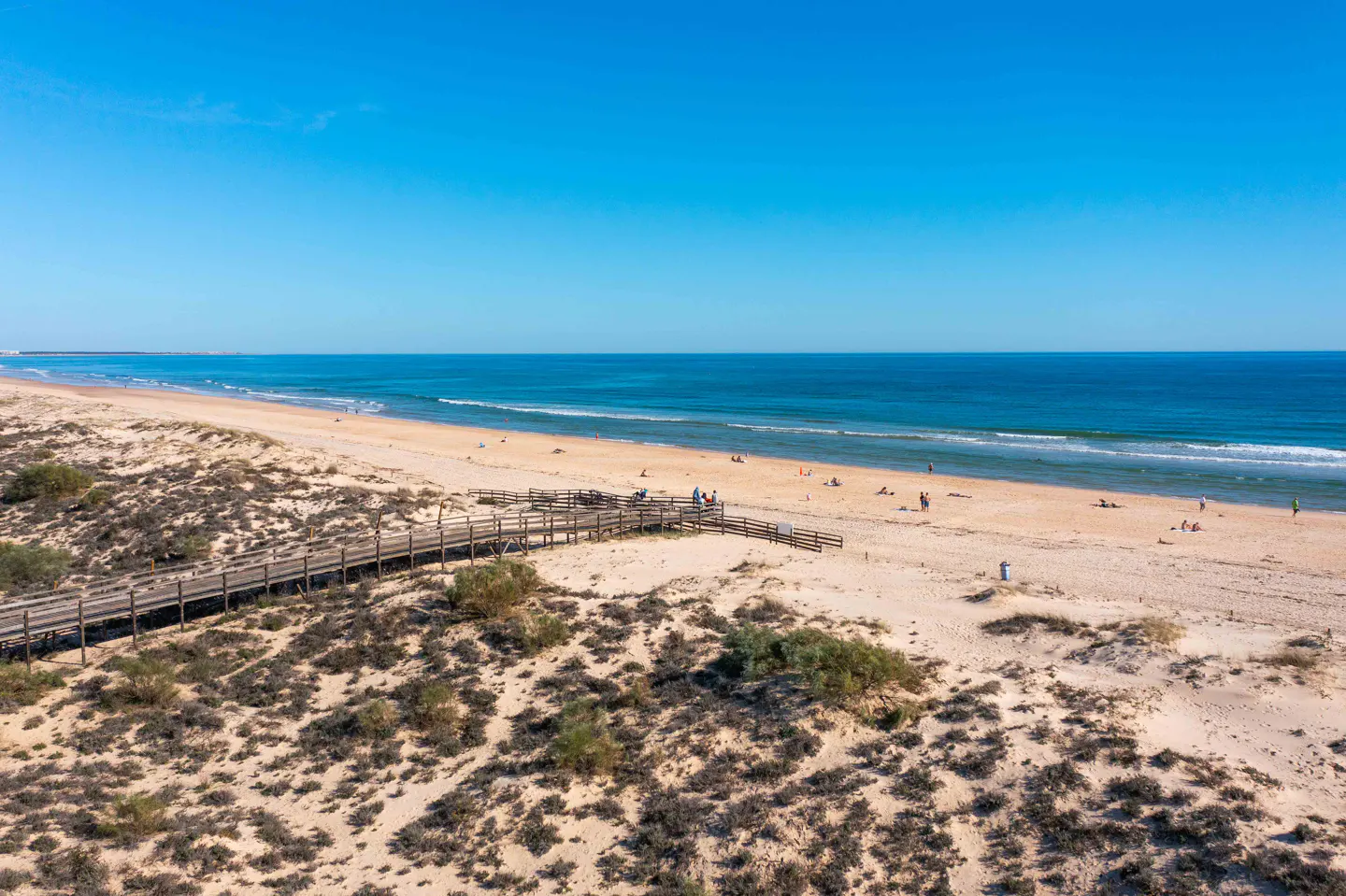 Beach scene with a wooden boardwalk leading to the sandy shore, blue ocean, and clear sky. People are scattered along the beach.