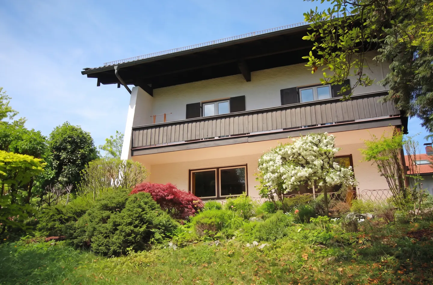 Two-story house with a brown balcony and black shutters, surrounded by lush green landscaping.