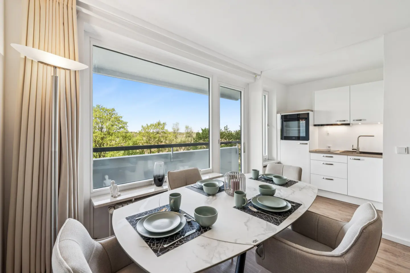 Bright apartment dining area with a white marble table set for six, overlooking a balcony and green trees. White kitchen in the background.
