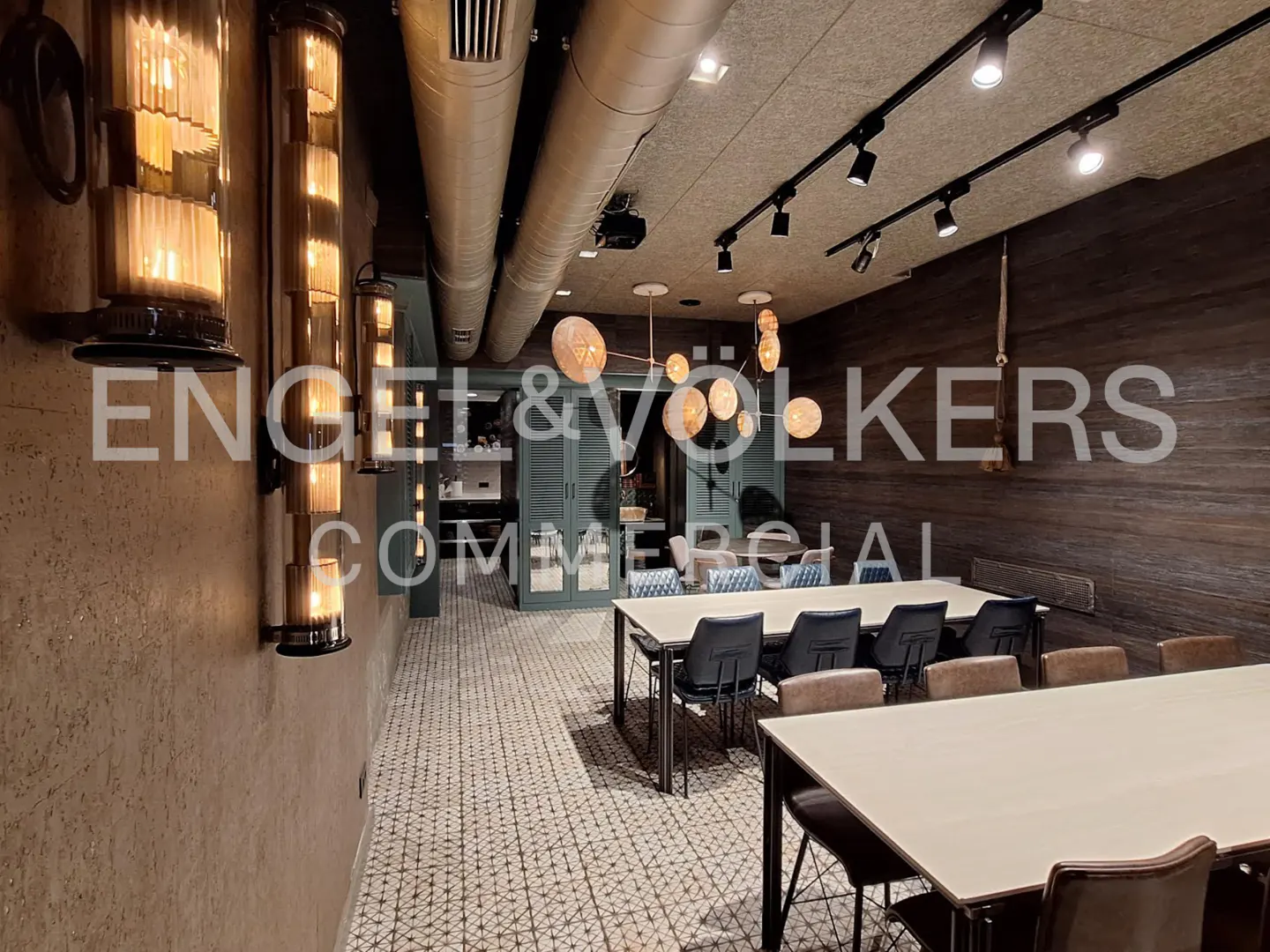 Office space with tables, chairs, and unique lighting fixtures. Exposed pipes and track lighting on the ceiling. Neutral color palette.