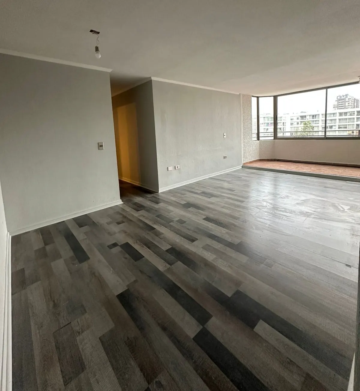 Empty living room with grey wood-look flooring, light grey walls, and a large window overlooking a city building.