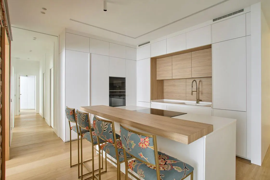 A modern kitchen with white cabinets, a light wood island, and four floral-patterned bar stools with gold frames. A hallway is visible in the background.