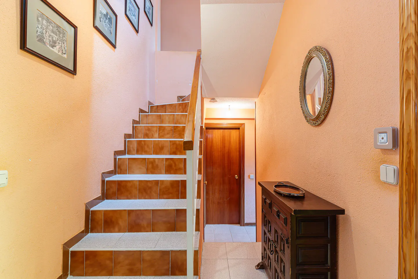 Interior view of a peach-colored hallway with a staircase, wooden door, dresser, and framed pictures.